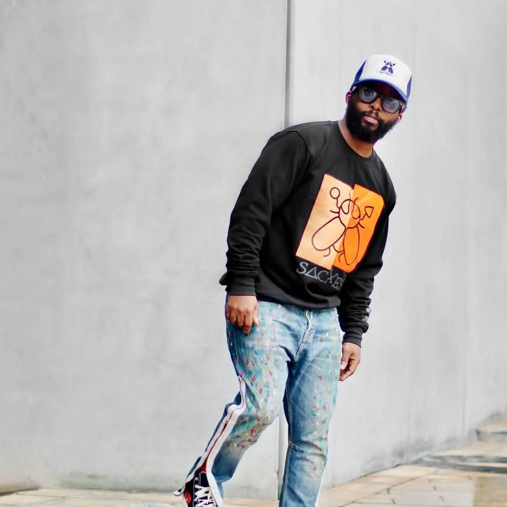 A man in a SACXESS trucker cap and black beetle-print sweatshirt walking past a pale wall